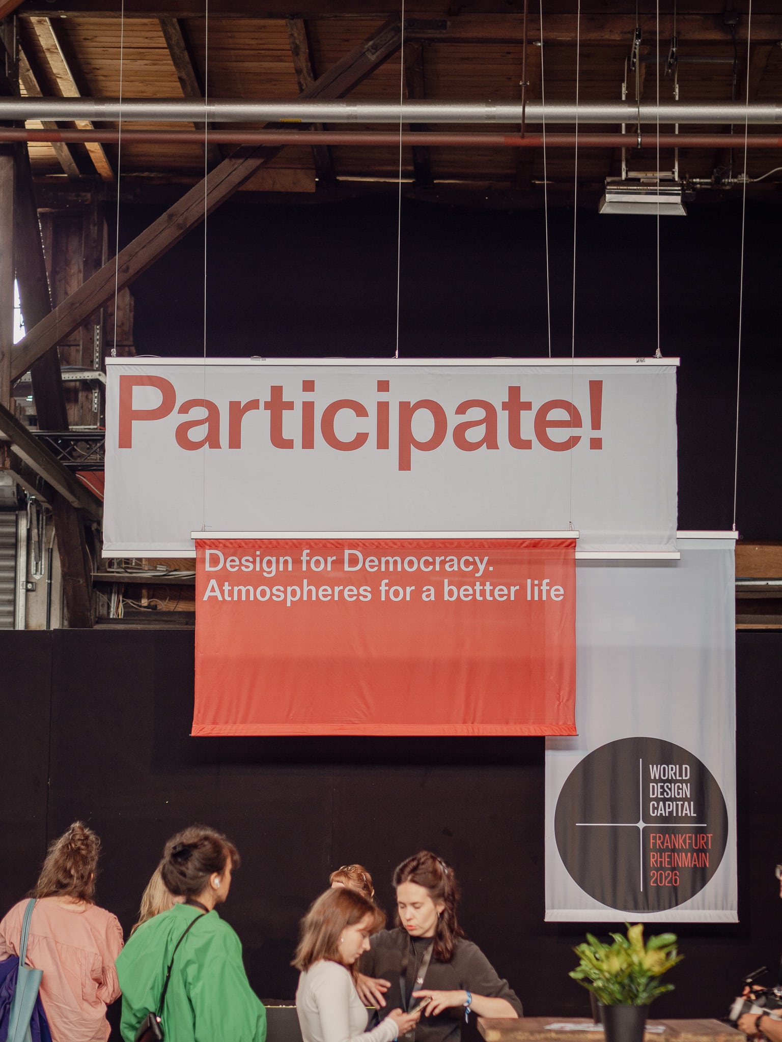 Menschen versammeln sich unter großen Bannern mit der Aufschrift "Participate! Design for Democracy. Atmospheres for a better life" in einer Veranstaltungshalle.
