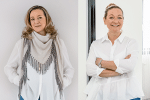 Sabine Kraus und Katharina Kraus stehen nebeneinander, beide tragen weiße Hemden und blaue Jeans. Sabine trägt zusätzlich einen grauen Fransenschal. Beide lächeln in die Kamera.