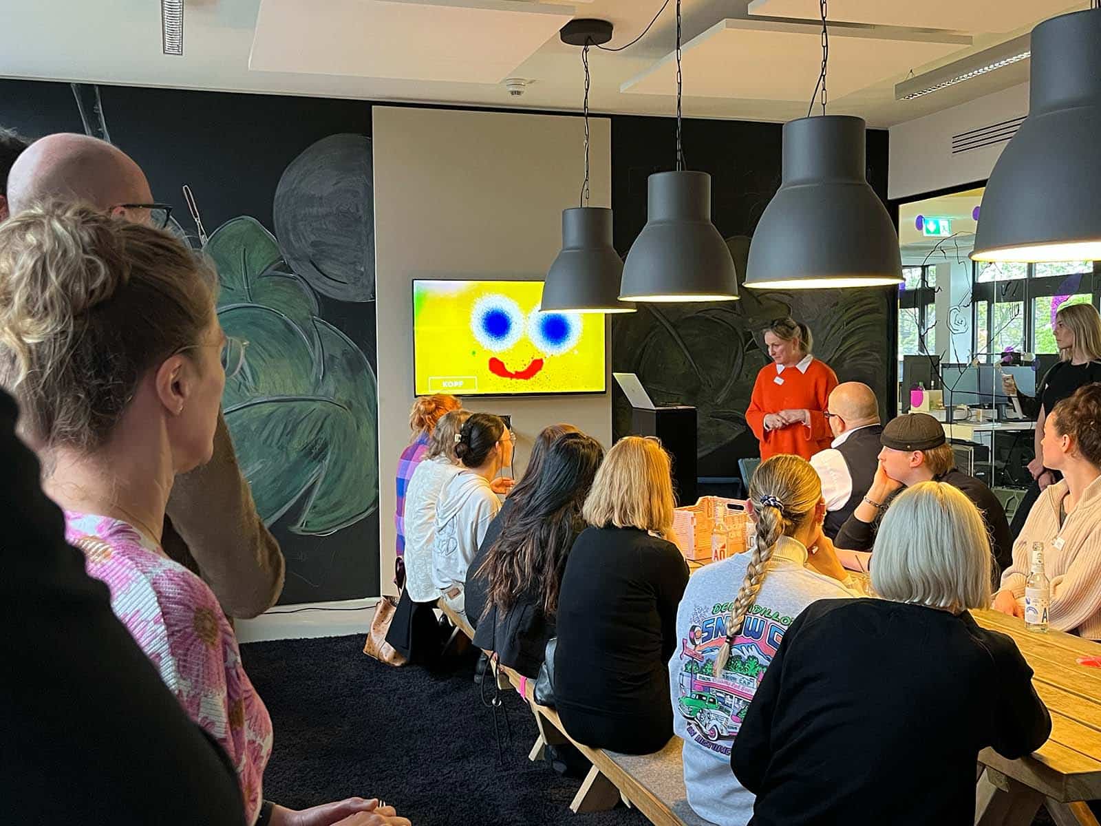 Eine Gruppe von Menschen sitzt an einem langen Tisch und hört einer Präsentation zu, die auf einem Bildschirm mit einem lächelnden gelben Smiley gezeigt wird. Sabine Kraus steht vorne und hält die Präsentation.