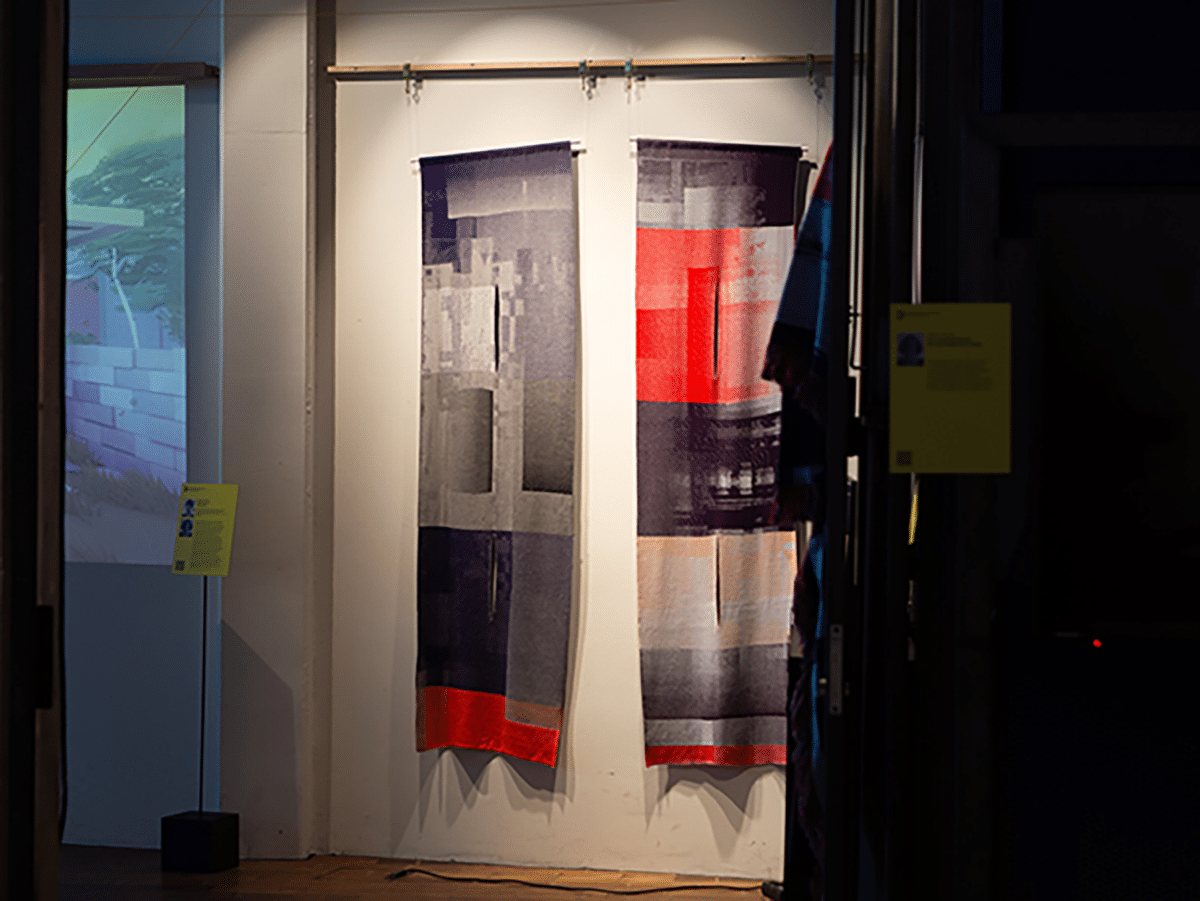 Zwei hängende Wandteppiche mit abstrakten Mustern in Rot-, Grau- und Lilatönen. Im Hintergrund ist eine Projektion auf einer Wand und eine Informationstafel zu sehen.