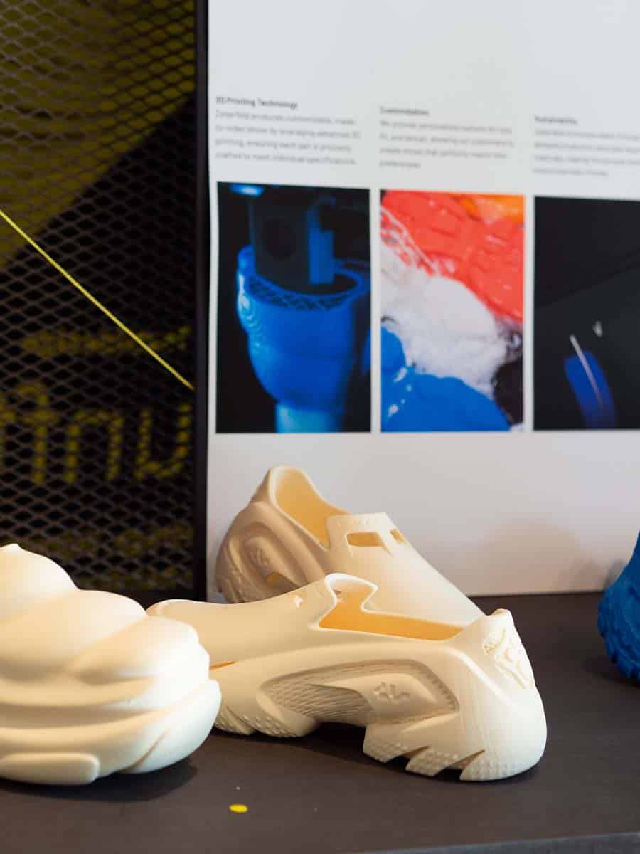 Zwei weiße, futuristische Schuhe aus 3D-Druck auf einem Tisch. Im Hintergrund sind Poster mit Informationen zu 3D-Drucktechnologien zu sehen.