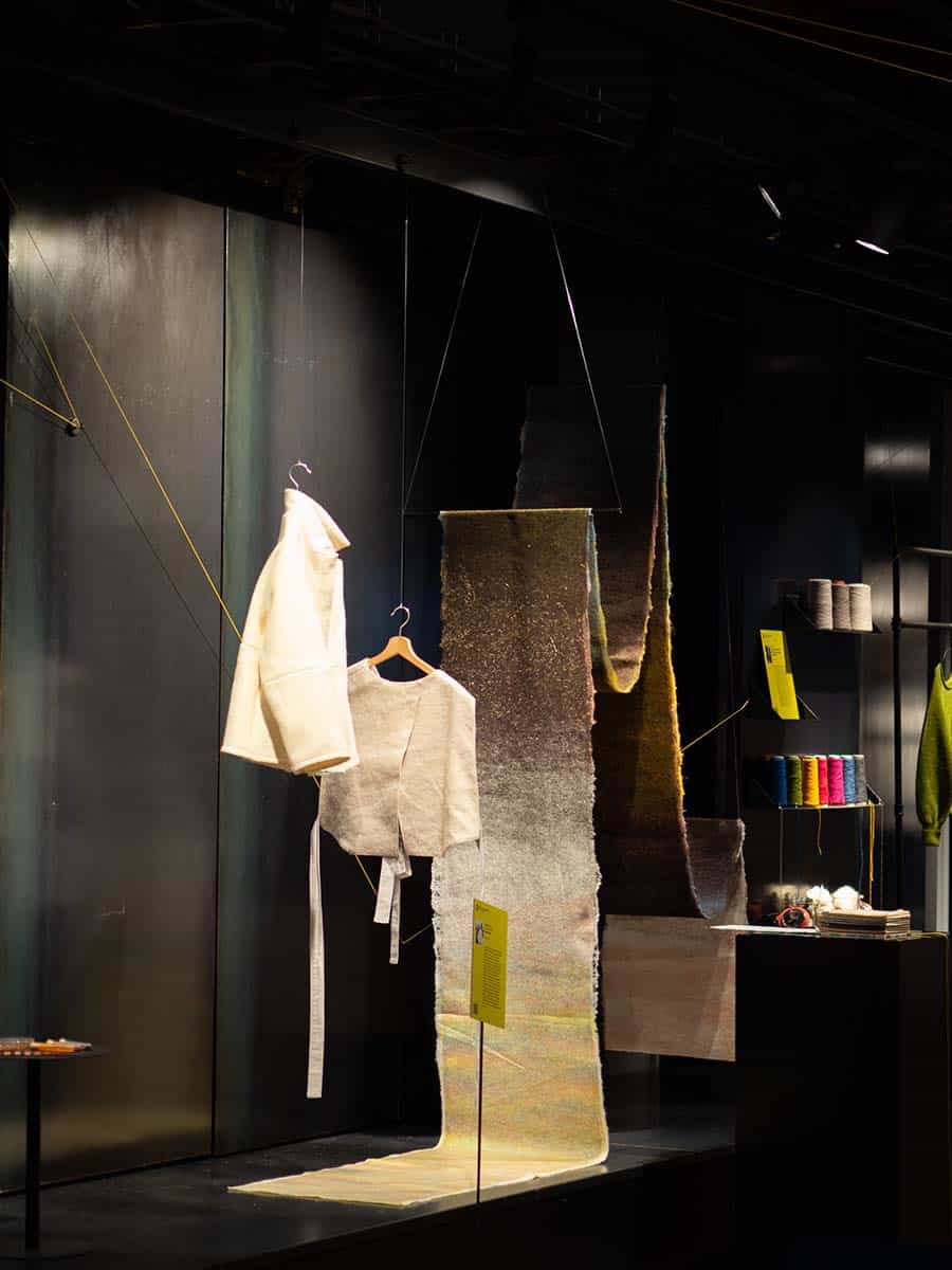 Ein Ausstellungsstück, das weiße, strukturierte Kleidung auf Kleiderbügeln zeigt, die von der Decke hängen. Weitere ausgestellte Textilien und Garnrollen sind im Hintergrund.