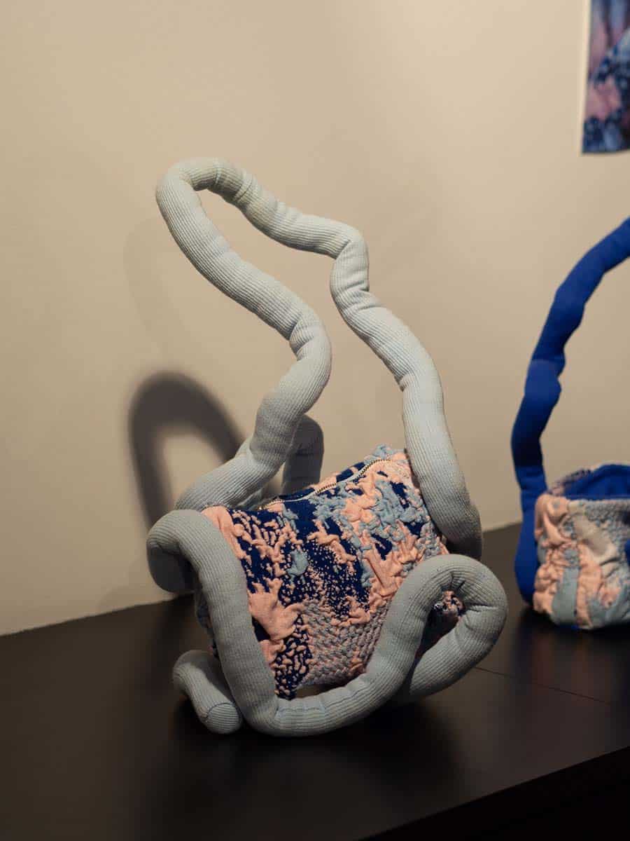 Eine skulpturale Handtasche mit blauen und rosa Mustern. Sie ist von einem flexiblen, blauen Griff umschlungen, der in geschwungenen Linien verläuft.