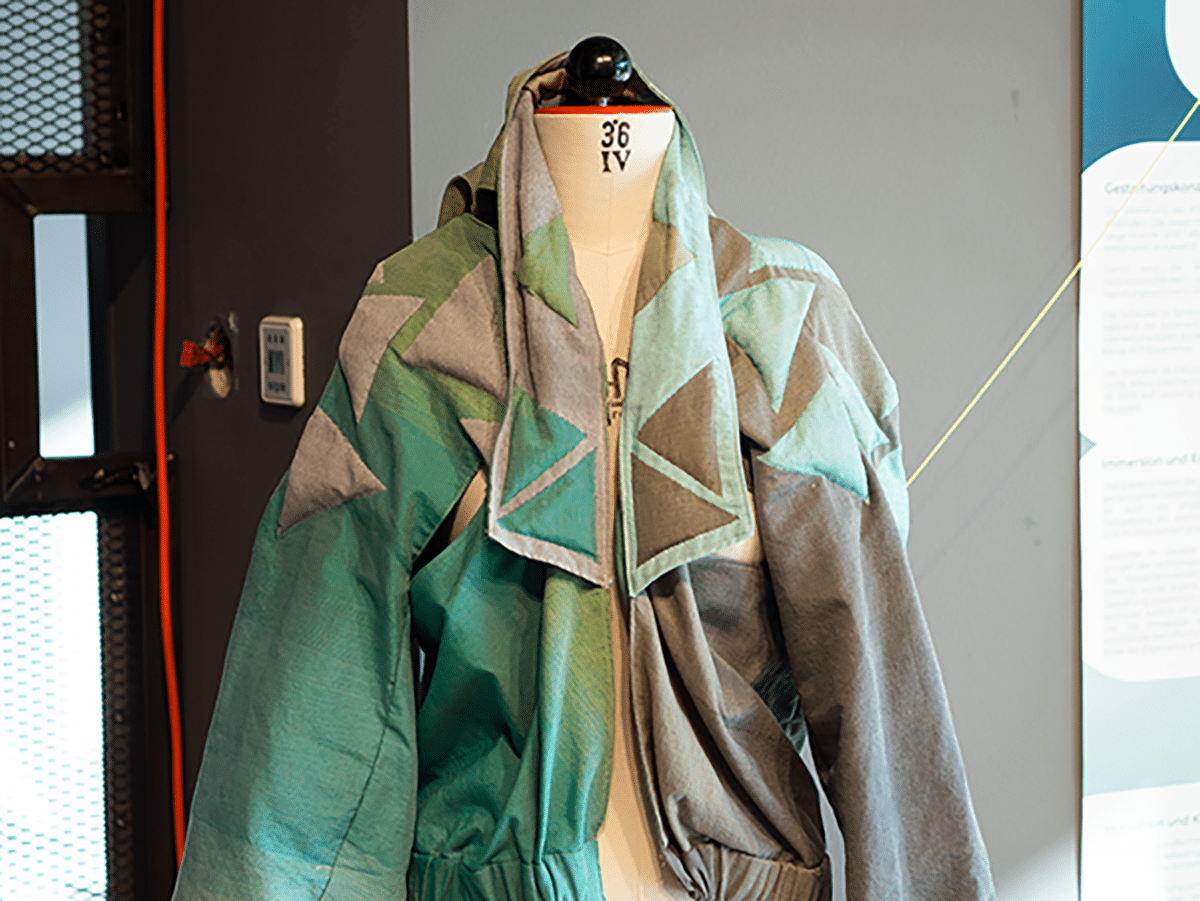 Eine Schneiderpuppe trägt eine Jacke in verschiedenen Grüntönen mit geometrischen Mustern. Im Hintergrund sind ein Thermostat und eine Informationstafel zu sehen.