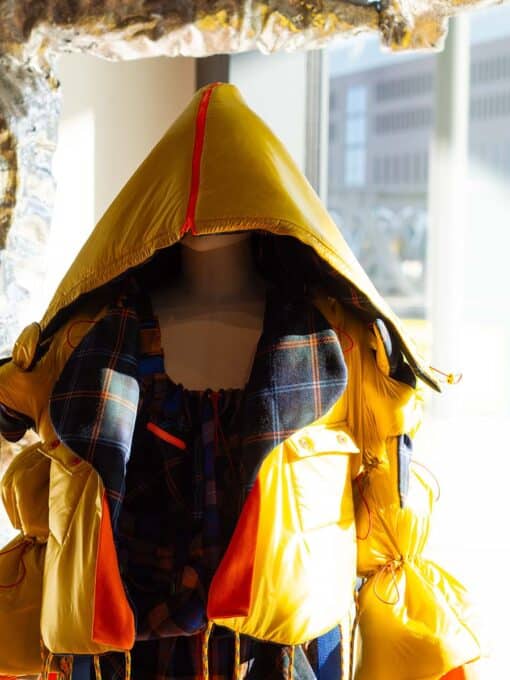 Eine Schneiderpuppe trägt eine gelbe, voluminöse Jacke mit Kapuze und roten Details. Darunter ist ein kariertes Kleidungsstück sichtbar. Im Hintergrund sind ein Fenster und eine skulpturale Struktur zu sehen.