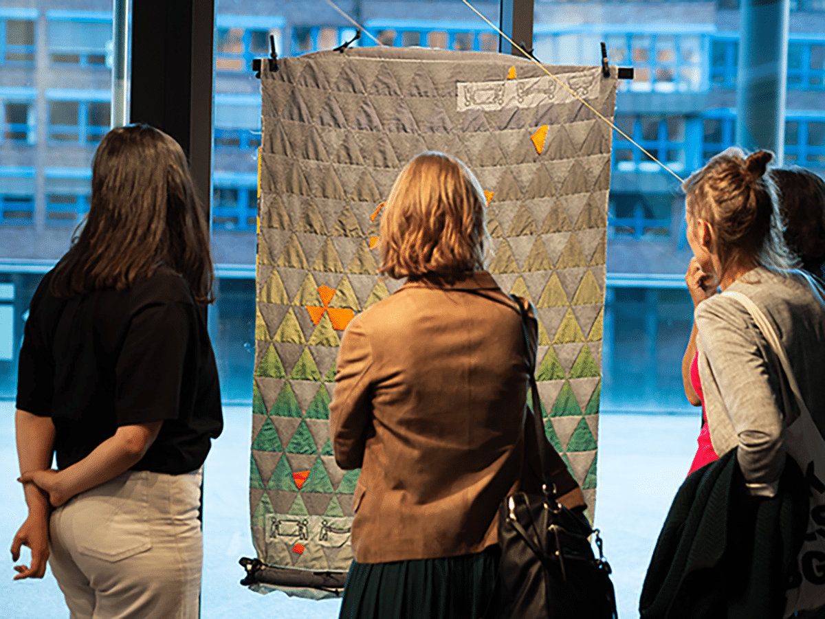 Vier Personen betrachten einen großen Wandteppich mit einem geometrischen Muster aus Dreiecken in Grau- und Grüntönen. Im Hintergrund sind Fenster mit Blick auf ein Gebäude.