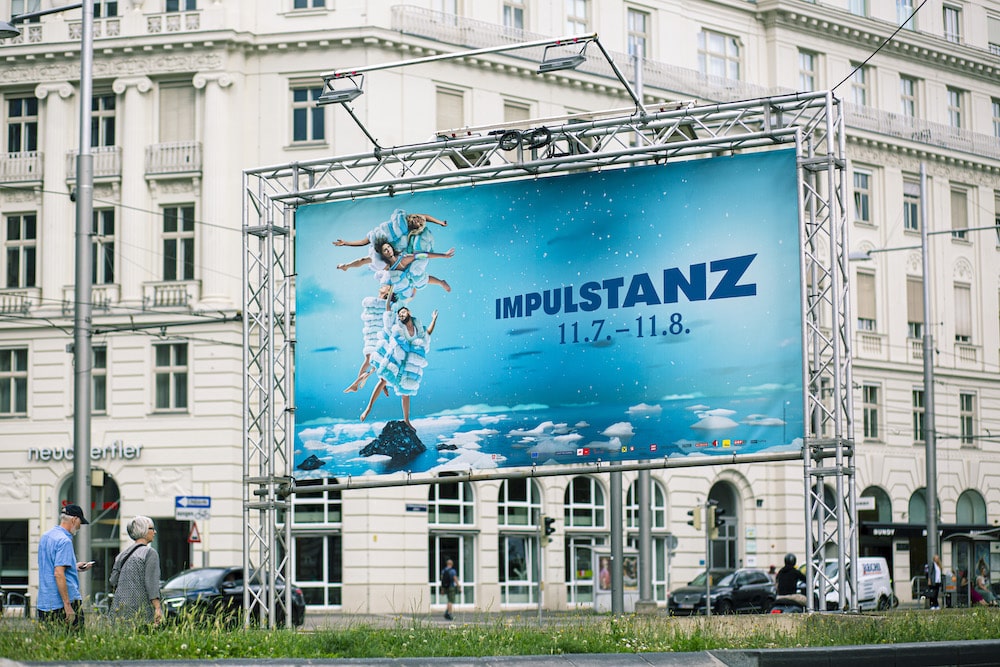 Kampagnen-Plakat für das Tanzfestivals ImPulsTanz, von der KI entwickeltes Motiv mit Menschen und Handtüchern mit einem Hintergrund aus Wolken und Sternenhimmel, blau-türkis