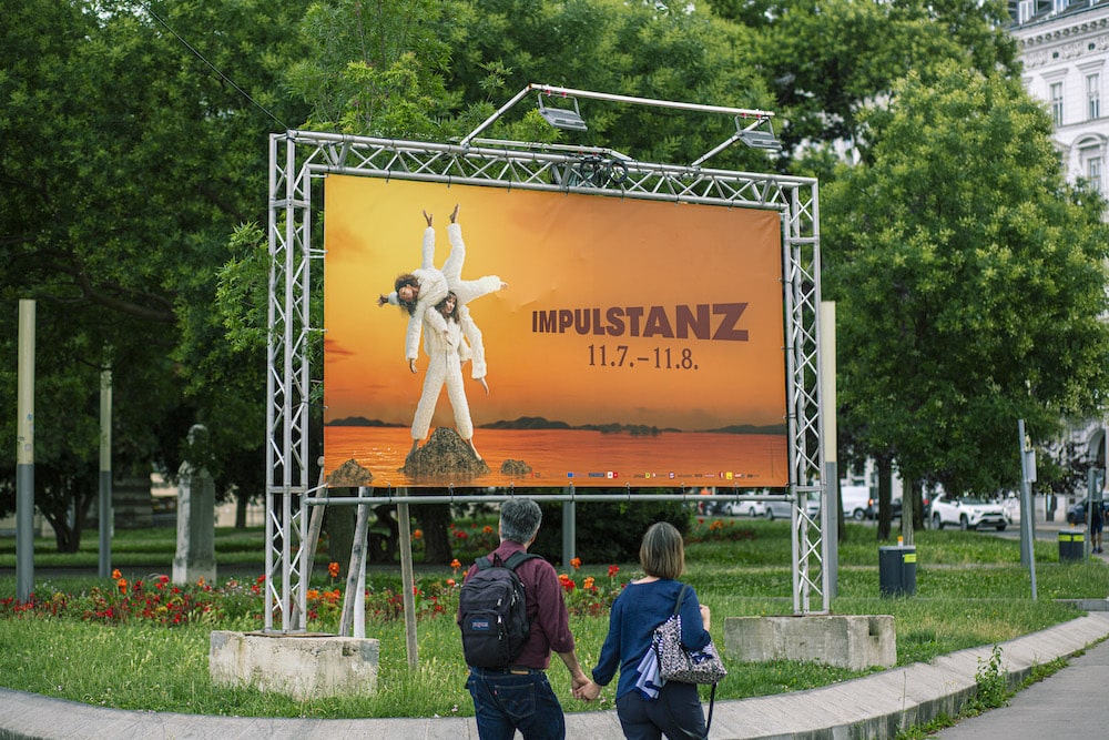 Kampagnen-Plakat für das Tanzfestivals ImPulsTanz, von der KI entwickeltes Motiv mit verknoteten Menschen in weißen Anzügen, orange-rot