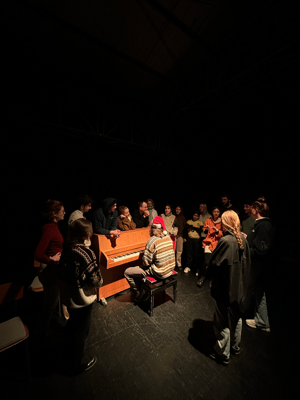 Eine Gruppe von Menschen steht im Kreis um ein Klavier. Eine Person mit einer Weihnachtmütze spielt das Klavier in einem dunklen Raum.