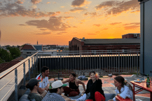 Eine Gruppe von Menschen sitzt und entspannt sich auf einem Dach, während die Sonne untergeht und den Himmel orange färbt.