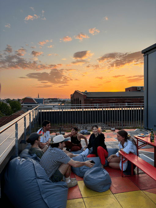 Eine Gruppe von Menschen sitzt und entspannt sich auf einem Dach, während die Sonne untergeht und den Himmel orange färbt.