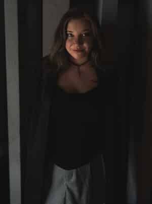 Lana Kari steht mit offenen Haaren in einem Türrahmen und schaut lächelnd in die Kamera. Die dunkle Lichtstimmung und warme Hauttöne lenken den Blick direkt auf ihr Gesicht