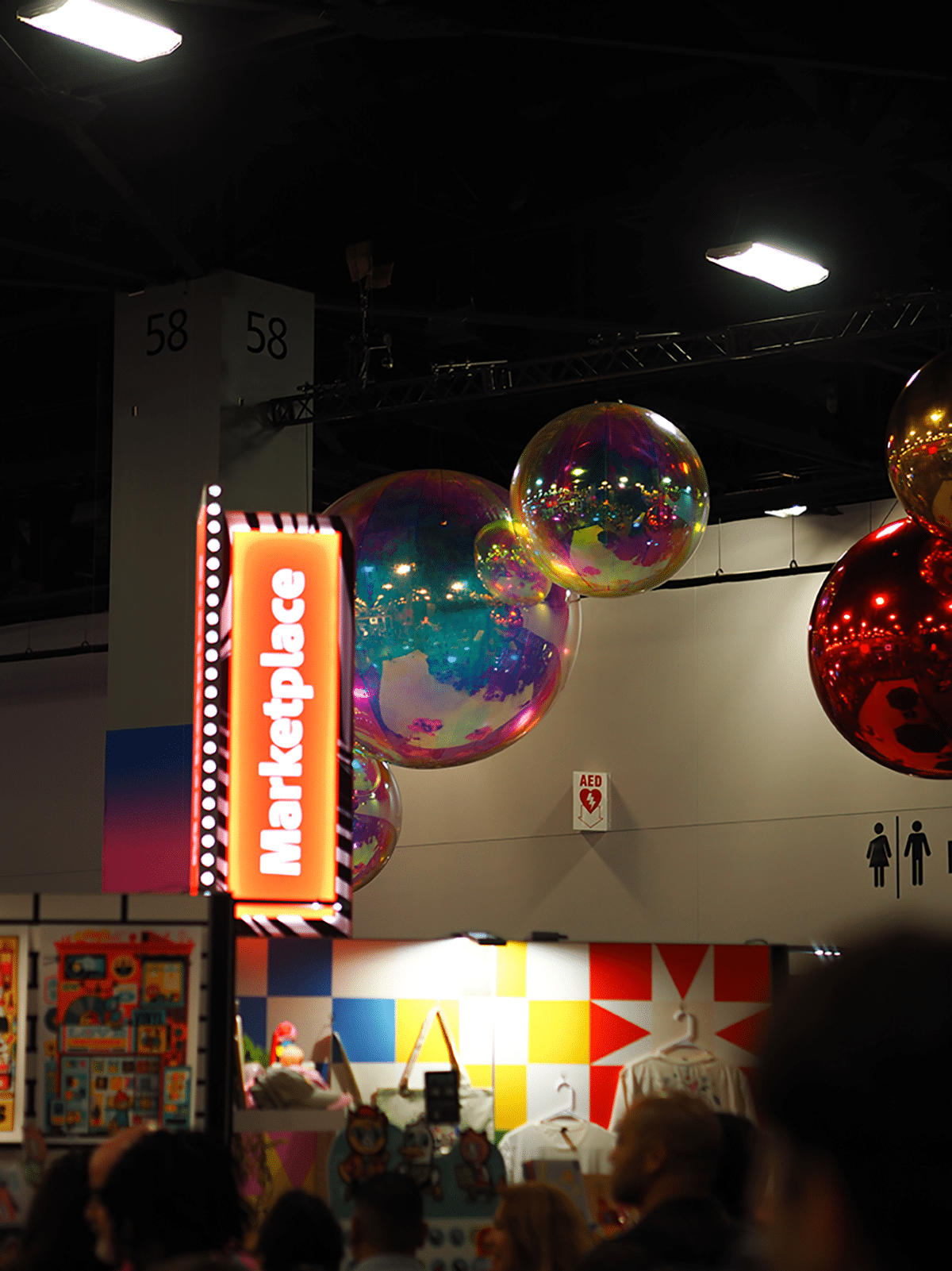 Ein Schild mit der Aufschrift "Marketplace" hängt im Vordergrund, während im Hintergrund eine Ausstellung mit bunten Produkten und schwebenden, reflektierenden Ballons zu sehen ist.