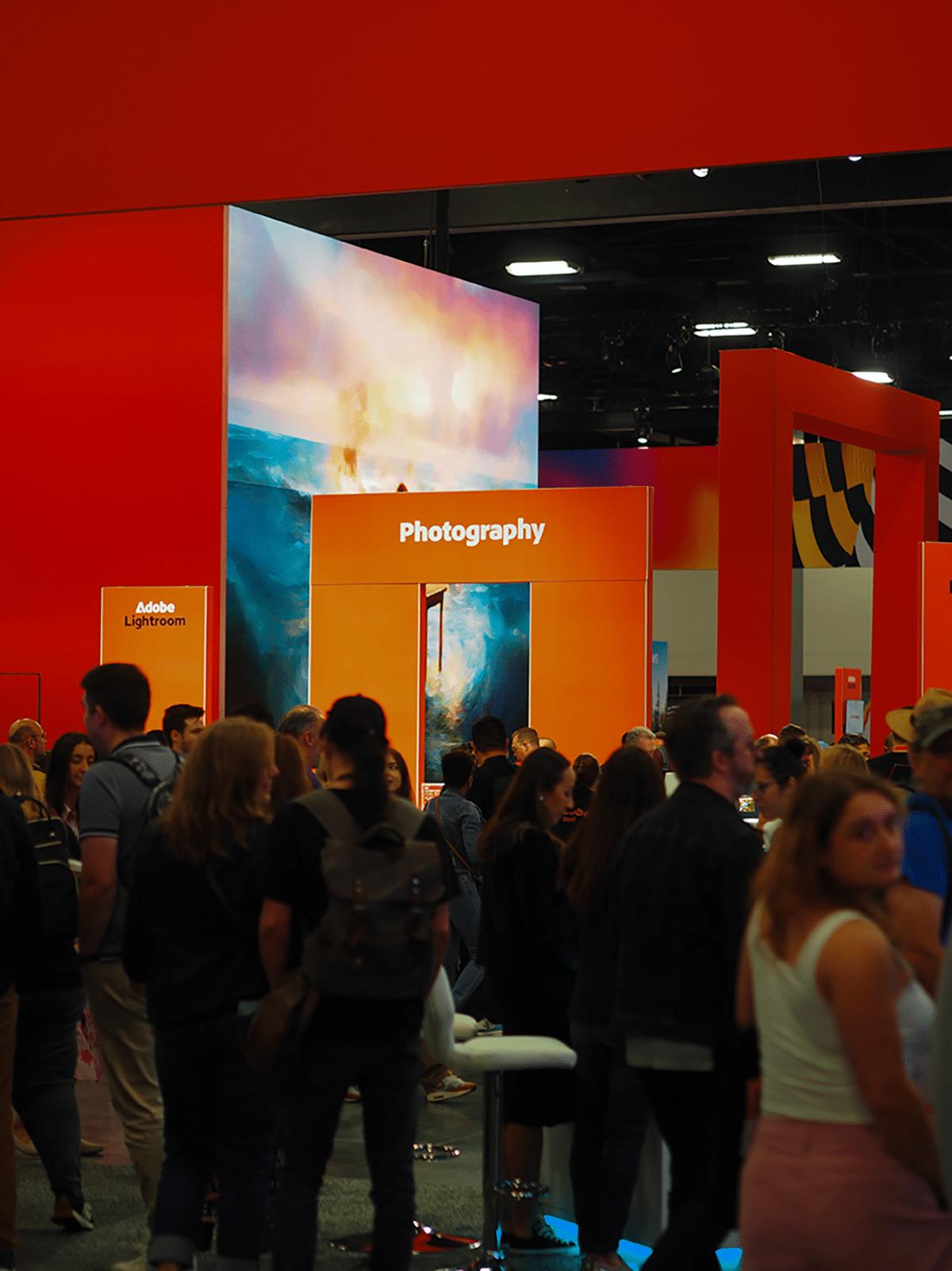 Eine Gruppe von Menschen steht vor einem orangefarbenen Stand mit der Aufschrift "Photography" bei der Adobe MAX Messe, umgeben von Ausstellungen und einem regen Besucherverkehr.