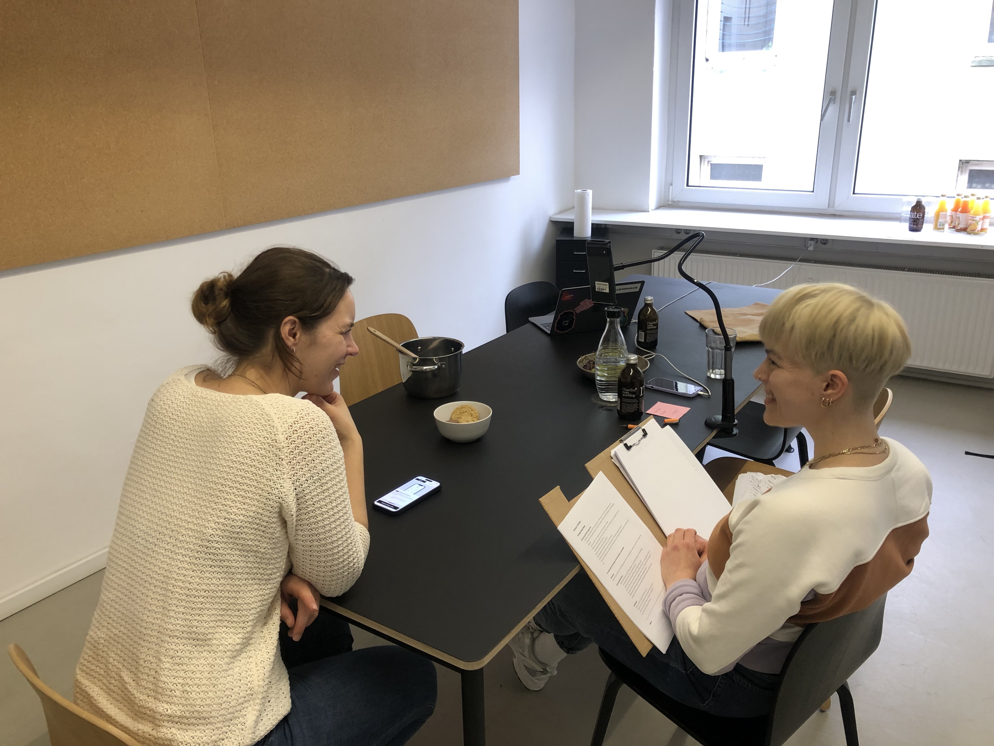 Zwei Personen sitzen an einem Tisch. Links testet eine Person eine App auf einem Smartphone, rechts notiert eine andere Person Beobachtungen auf einem Klemmbrett. Auf dem Tisch stehen Getränke, ein Laptop und ein Topf. Im Hintergrund ein Fenster.