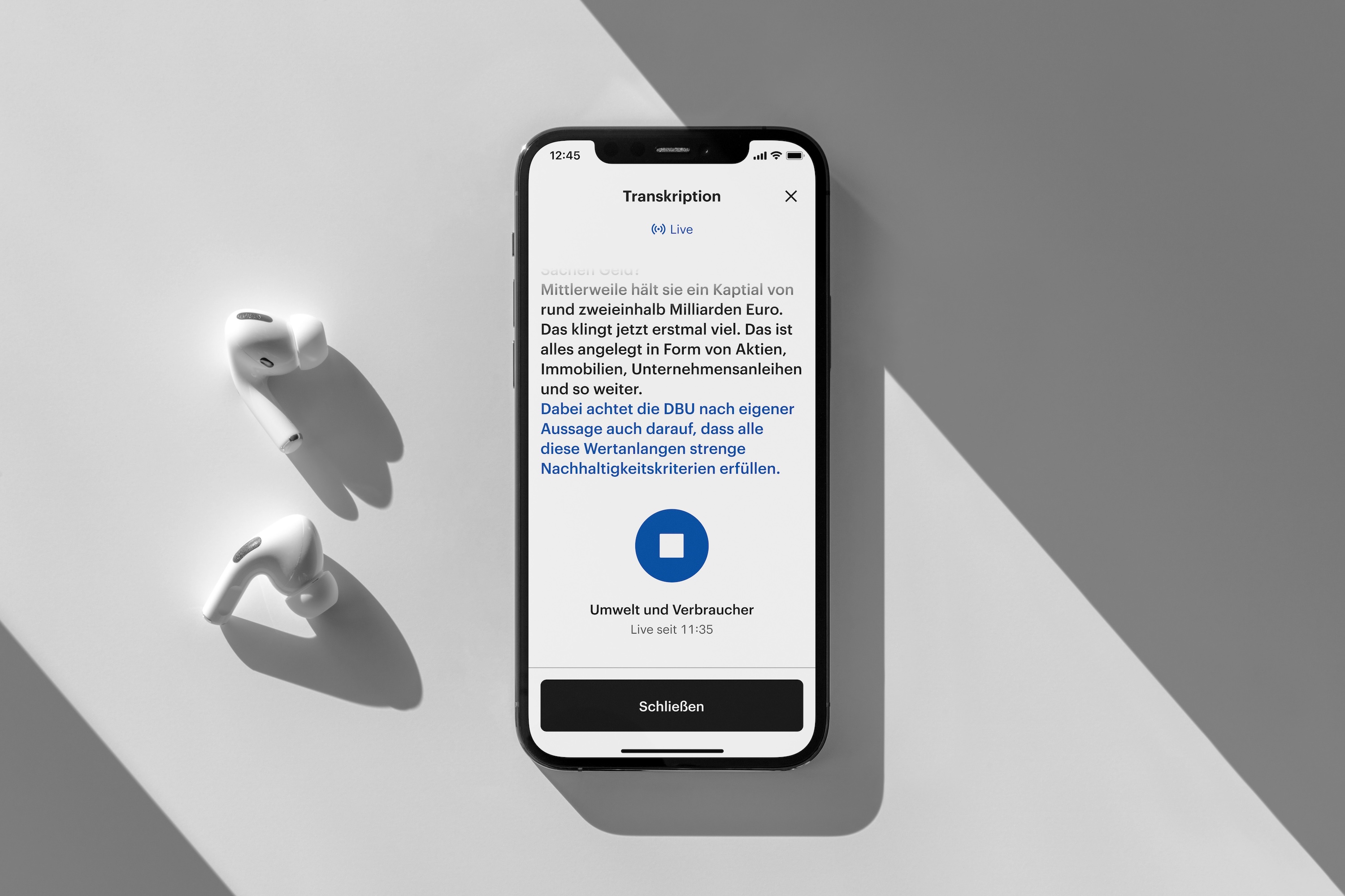 Smartphone auf hellem Hintergrund zeigt eine Live-Transkription von gesprochenem Text. Zwei kabellose Kopfhörer liegen daneben.
