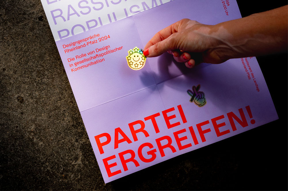 Agenturporträt: agentur alma, Designgespräche Rheinland-Pfalz 2024 – Corporate Design, Lila Plakat mit roter Schrift, eine Hand klebt einen gelben Smileysticker darauf