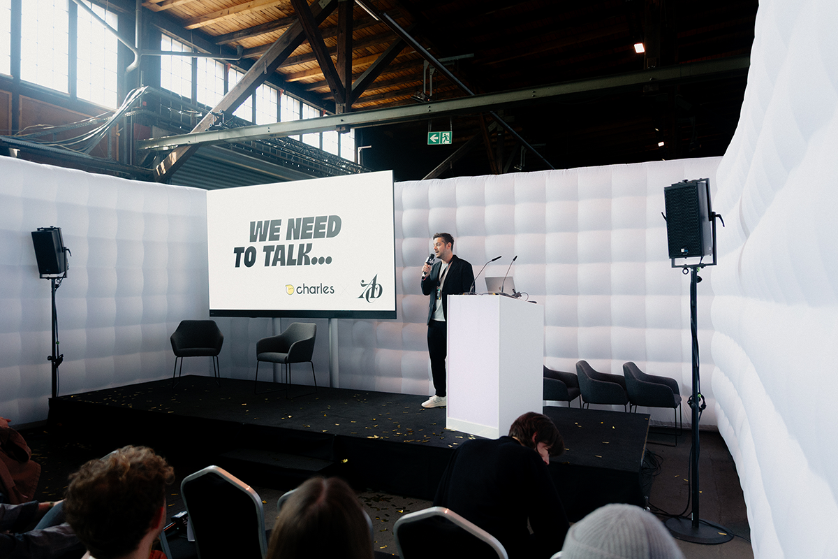 Speaker präsentiert auf kleiner Bühne vor weiß gepolsterten Wänden, eine Folie zeigt den Titel „WE NEED TO TALK…“ mit Logos von Charles und dem ADC.