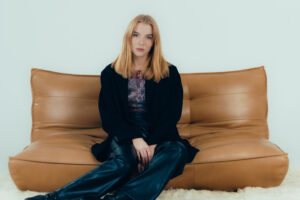 junge blonde frau in einem dunklem Outift, sitzend auf einer Ledercouch in einem weißen Studio.