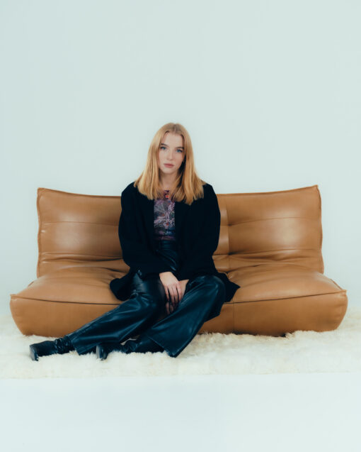 junge blonde frau in einem dunklem Outift, sitzend auf einer Ledercouch in einem weißen Studio.