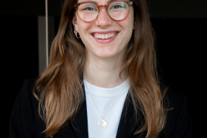 Junge Frau, Jana Reske, mit langen Haaren und Brille, die lächelnd in schwarzem Blazer und weißem Shirt in einem Innenraum in die Kamera schaut.