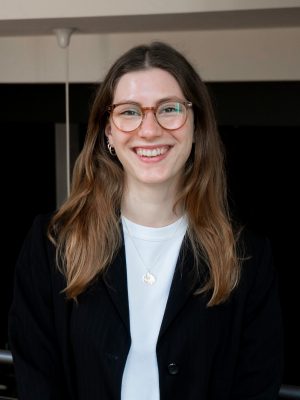 Junge Frau, Jana Reske, mit langen Haaren und Brille, die lächelnd in schwarzem Blazer und weißem Shirt in einem Innenraum in die Kamera schaut.