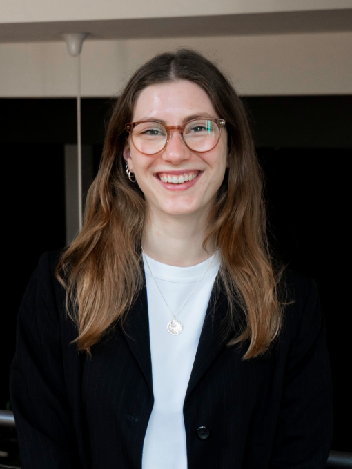 Junge Frau, Jana Reske, mit langen Haaren und Brille, die lächelnd in schwarzem Blazer und weißem Shirt in einem Innenraum in die Kamera schaut.
