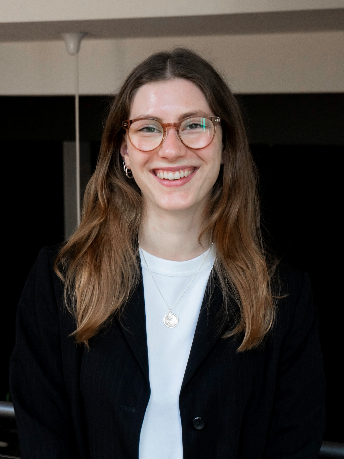 Junge Frau, Jana Reske, mit langen Haaren und Brille, die lächelnd in schwarzem Blazer und weißem Shirt in einem Innenraum in die Kamera schaut.