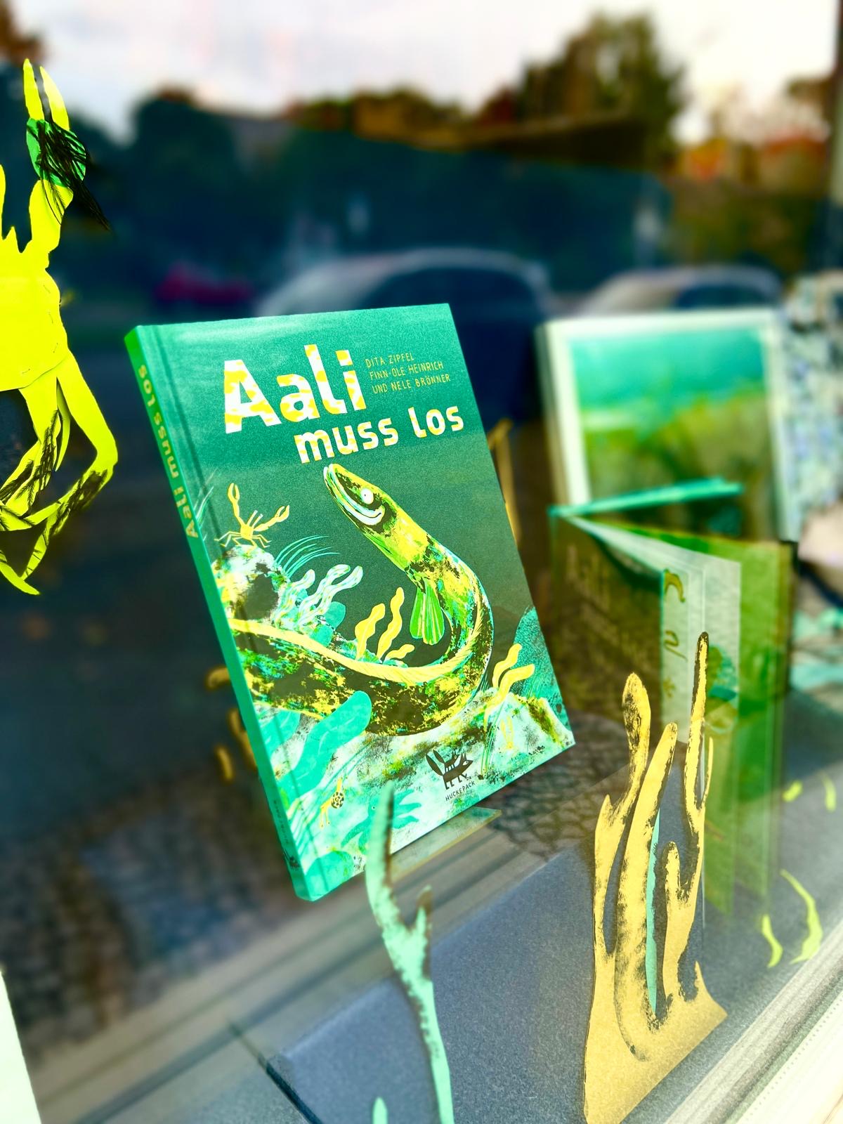 Fotografie eines Buches mit dem Titel „Aali muss los“ im Schaufenster. Das Cover zeigt einen gezeichneten Aal in einer grünen Unterwasserwelt. Spiegelungen von Straße und Bäumen sind im Glas sichtbar, daneben weitere Buchillustrationen und Dekoelemente in Meeresoptik.