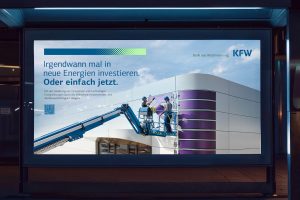 Eine nächtlich beleuchtete Werbetafel zeigt eine KfW-Anzeige. Links steht der Text „Irgendwann mal in neue Energien investieren. Oder einfach jetzt.“ Daneben arbeiten zwei Personen mit Schutzhelmen auf einer Hebebühne an der Fassade eines modernen Gebäudes mit violetten Paneelen. Der Himmel ist blau, oben rechts das KfW-Logo.
