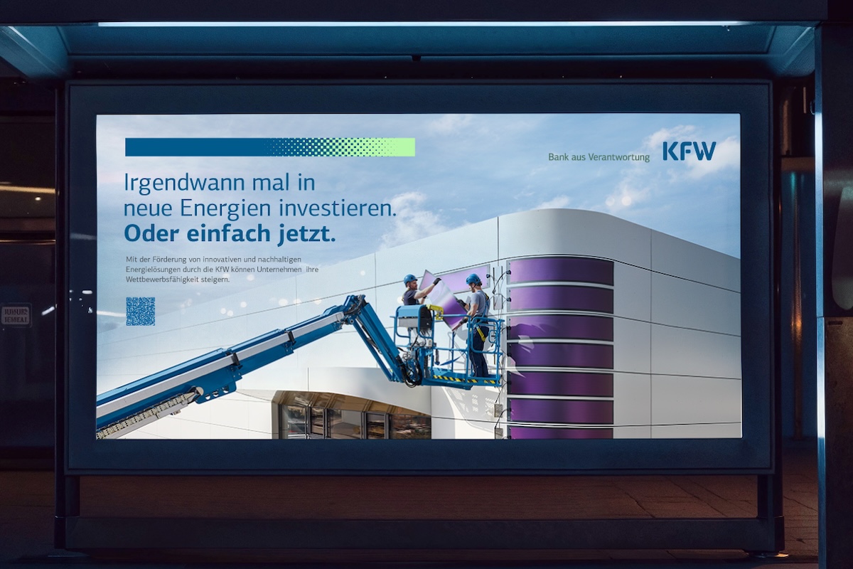 Eine nächtlich beleuchtete Werbetafel zeigt eine KfW-Anzeige. Links steht der Text „Irgendwann mal in neue Energien investieren. Oder einfach jetzt.“ Daneben arbeiten zwei Personen mit Schutzhelmen auf einer Hebebühne an der Fassade eines modernen Gebäudes mit violetten Paneelen. Der Himmel ist blau, oben rechts das KfW-Logo.