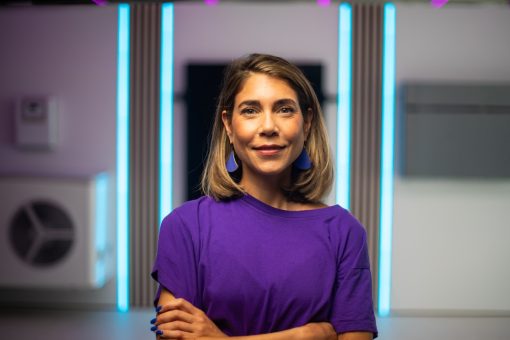 Eine Frau, Sophia Rödiger, mit schulterlangem, blond-braunem Haar steht in einem modern beleuchteten Innenraum. Sie trägt ein violettes T-Shirt und passende blaue Statement-Ohrringe. Mit leicht verschränkten Armen blickt sie freundlich und selbstbewusst in die Kamera. Hinter ihr leuchten vertikale LED-Linien in Blau und Magenta und erzeugen eine futuristische Atmosphäre.