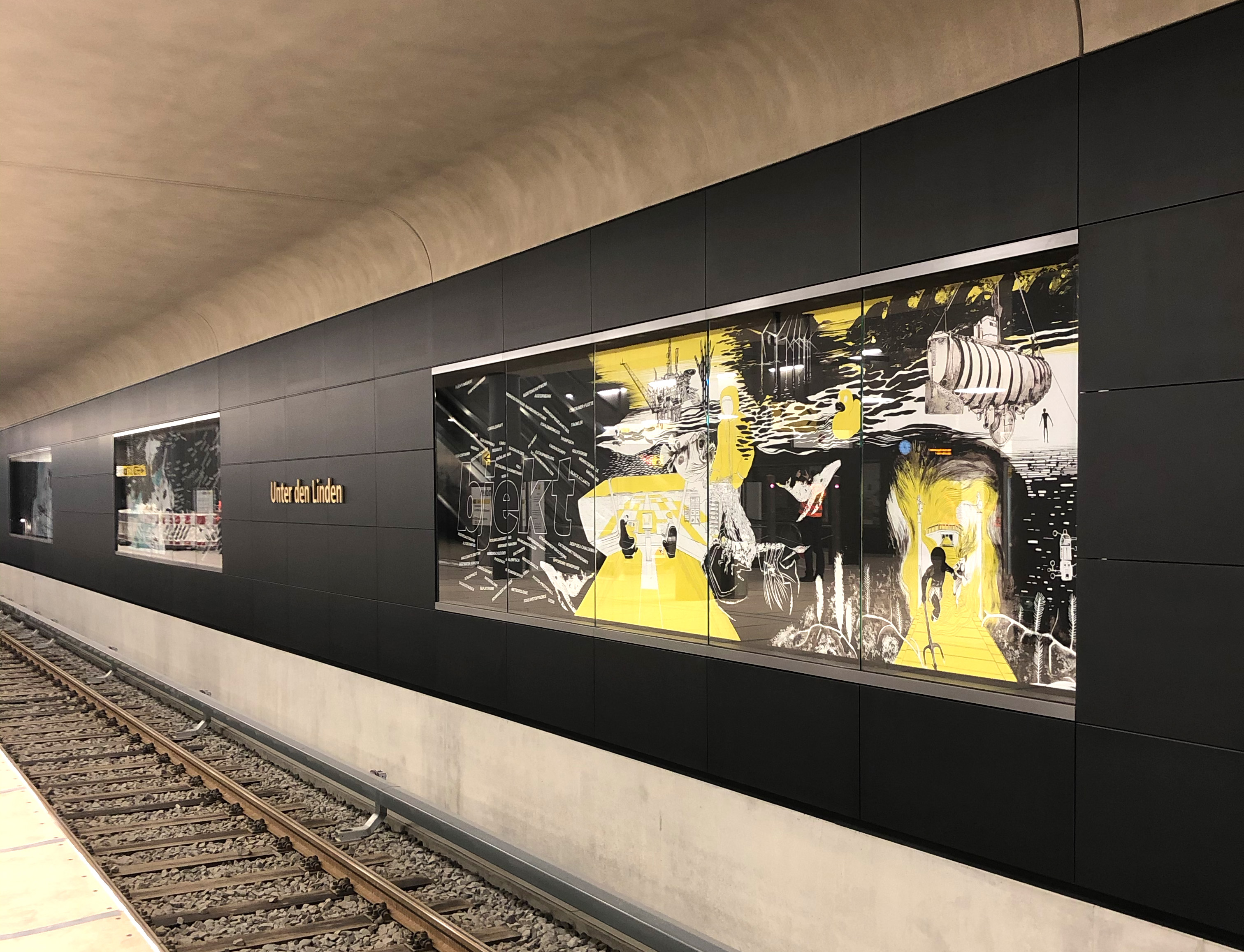 Eine Fotografie von mehreren Illustrationen, die eine Sammlung in der U Bahn Station Unter den Linden bilden. Die Illustrationen sind in schwarz, weiß und gelb gehalten und zeigen nautische, surreale Erzählungen von einem Schiff, einem Wal und einer Person mit Neptuns Dreizack