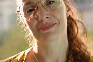 Porträtaufnahme einer Frau mit großer Brille und dezentem Lächeln, aufgenommen im weichen Gegenlicht vor unscharfem, urbanem Hintergrund. Sie trägt ein gelb-gemustertes Oberteil und eine filigrane Kette mit zwei ineinander verschlungenen Ringen.