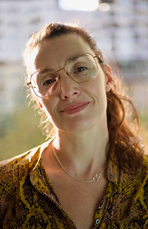 Porträtaufnahme einer Frau mit großer Brille und dezentem Lächeln, aufgenommen im weichen Gegenlicht vor unscharfem, urbanem Hintergrund. Sie trägt ein gelb-gemustertes Oberteil und eine filigrane Kette mit zwei ineinander verschlungenen Ringen.