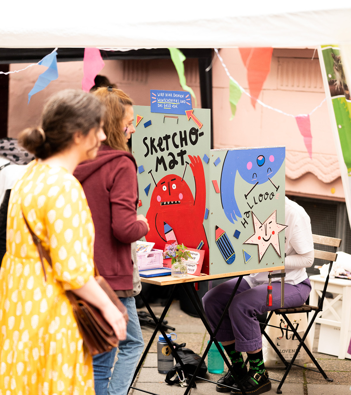 Eine Besucherin steht vor einem Stand mit bunten, handgezeichneten Figuren und dem Schriftzug „Sketchomat“. Eine Künstlerin sitzt dahinter und zeichnet.