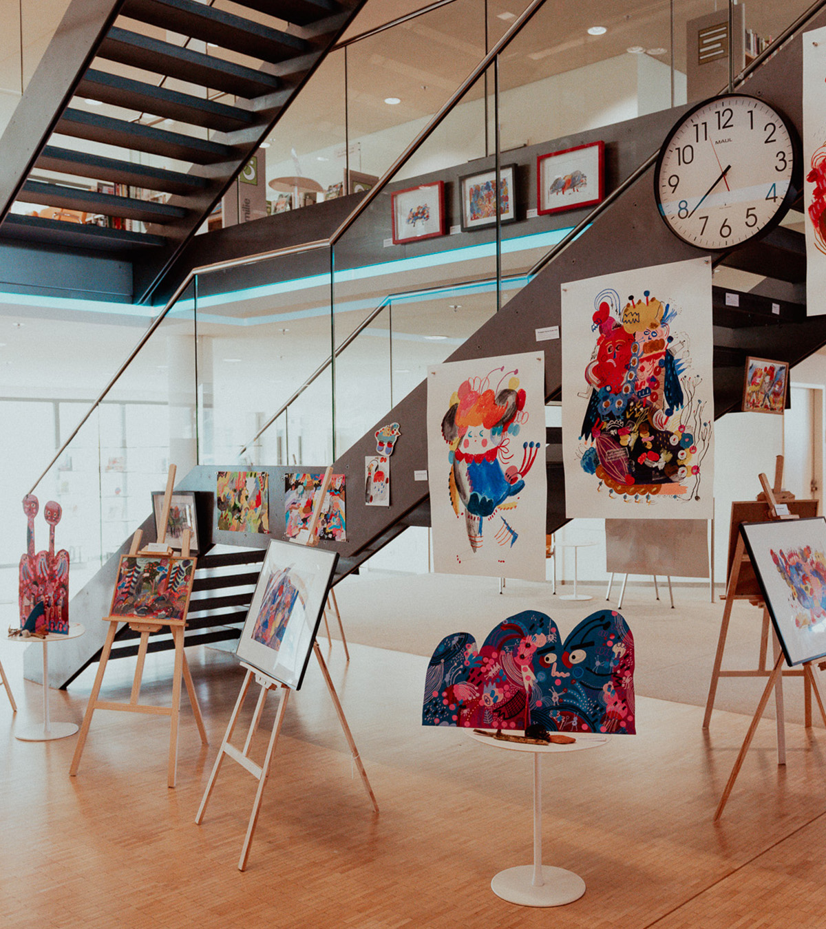 Mehrere farbige Kinderbuch-Illustrationen hängen an Staffeleien und Geländern in einem modernen, hellen Gebäude mit Glas und Holz.