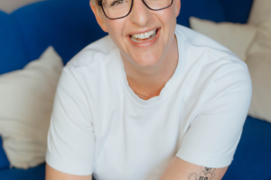 Eine kurzhaarige Person mit blondem Haar und schwarzer Brille sitzt lächelnd auf einem blauen Sofa. Sie trägt ein weißes T-Shirt und blickt offen in die Kamera; der Hintergrund ist weich unscharf mit hellen Kissen, die eine warme, entspannte Atmosphäre erzeugen.