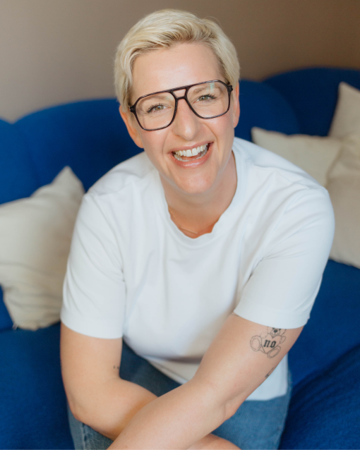 Eine kurzhaarige Person mit blondem Haar und schwarzer Brille sitzt lächelnd auf einem blauen Sofa. Sie trägt ein weißes T-Shirt und blickt offen in die Kamera; der Hintergrund ist weich unscharf mit hellen Kissen, die eine warme, entspannte Atmosphäre erzeugen.