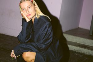 Junge Frau mit blondem Haar und bunten Haarspangen sitzt nachdenklich in Hocke auf Steinboden, trägt einen dunklen Oversize-Blazer und pinke Plateau-Schuhe; im Hintergrund eine Wand mit dem Schriftzug „Kultur“.