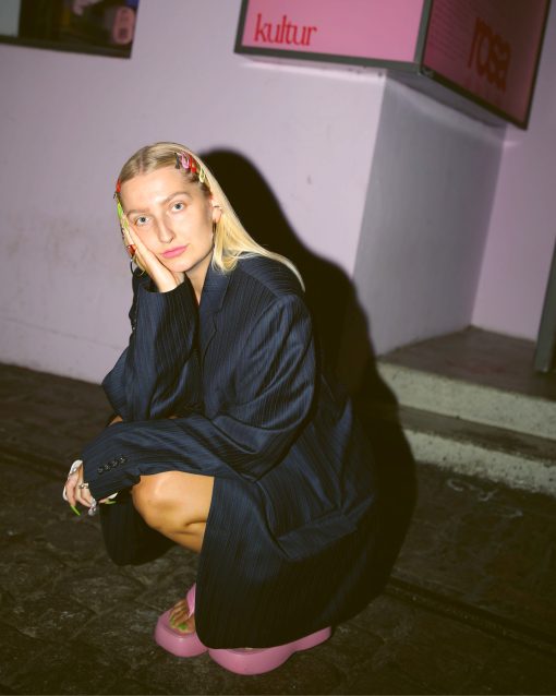 Junge Frau mit blondem Haar und bunten Haarspangen sitzt nachdenklich in Hocke auf Steinboden, trägt einen dunklen Oversize-Blazer und pinke Plateau-Schuhe; im Hintergrund eine Wand mit dem Schriftzug „Kultur“.