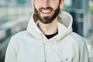 Porträt einer lächelnden Person mit Bart und zurückgebundenen Haaren, die einen hellen Hoodie trägt und in einem modernen, lichtdurchfluteten Glasgang steht.