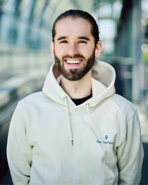 Porträt einer lächelnden Person mit Bart und zurückgebundenen Haaren, die einen hellen Hoodie trägt und in einem modernen, lichtdurchfluteten Glasgang steht.