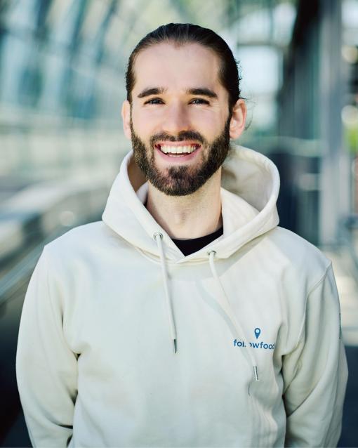 Porträt einer lächelnden Person mit Bart und zurückgebundenen Haaren, die einen hellen Hoodie trägt und in einem modernen, lichtdurchfluteten Glasgang steht.