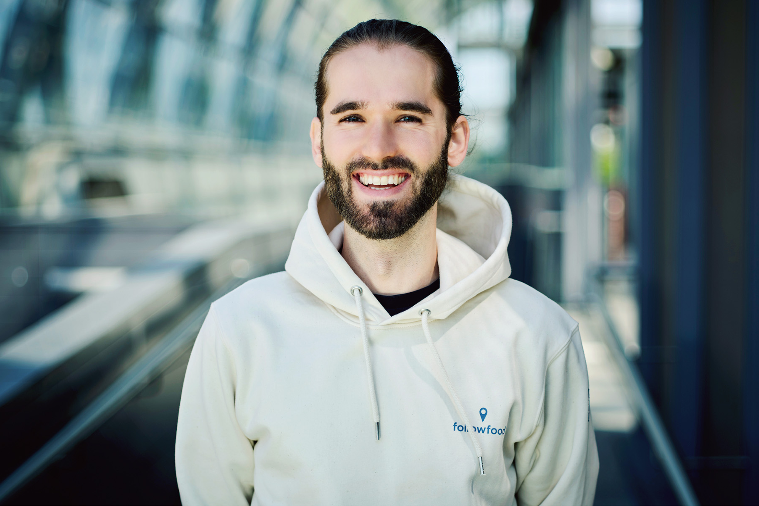 Porträt einer lächelnden Person mit Bart und zurückgebundenen Haaren, die einen hellen Hoodie trägt und in einem modernen, lichtdurchfluteten Glasgang steht.