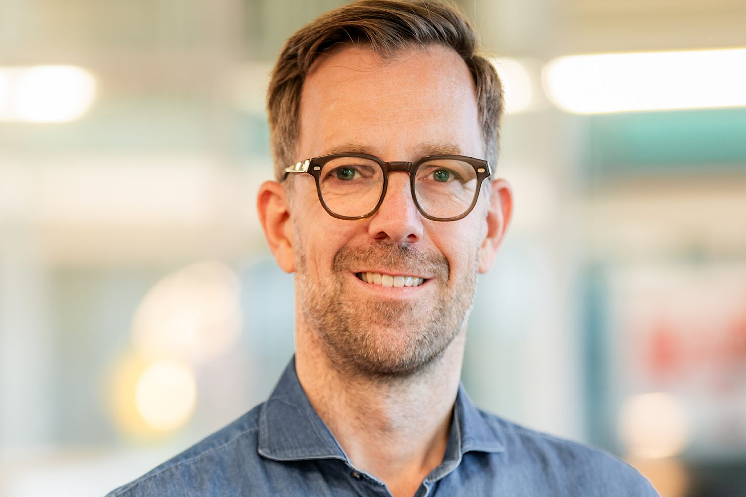 Porträt eines Mannes mit Brille und Bart, lächelnd vor unscharfem Bürohintergrund.