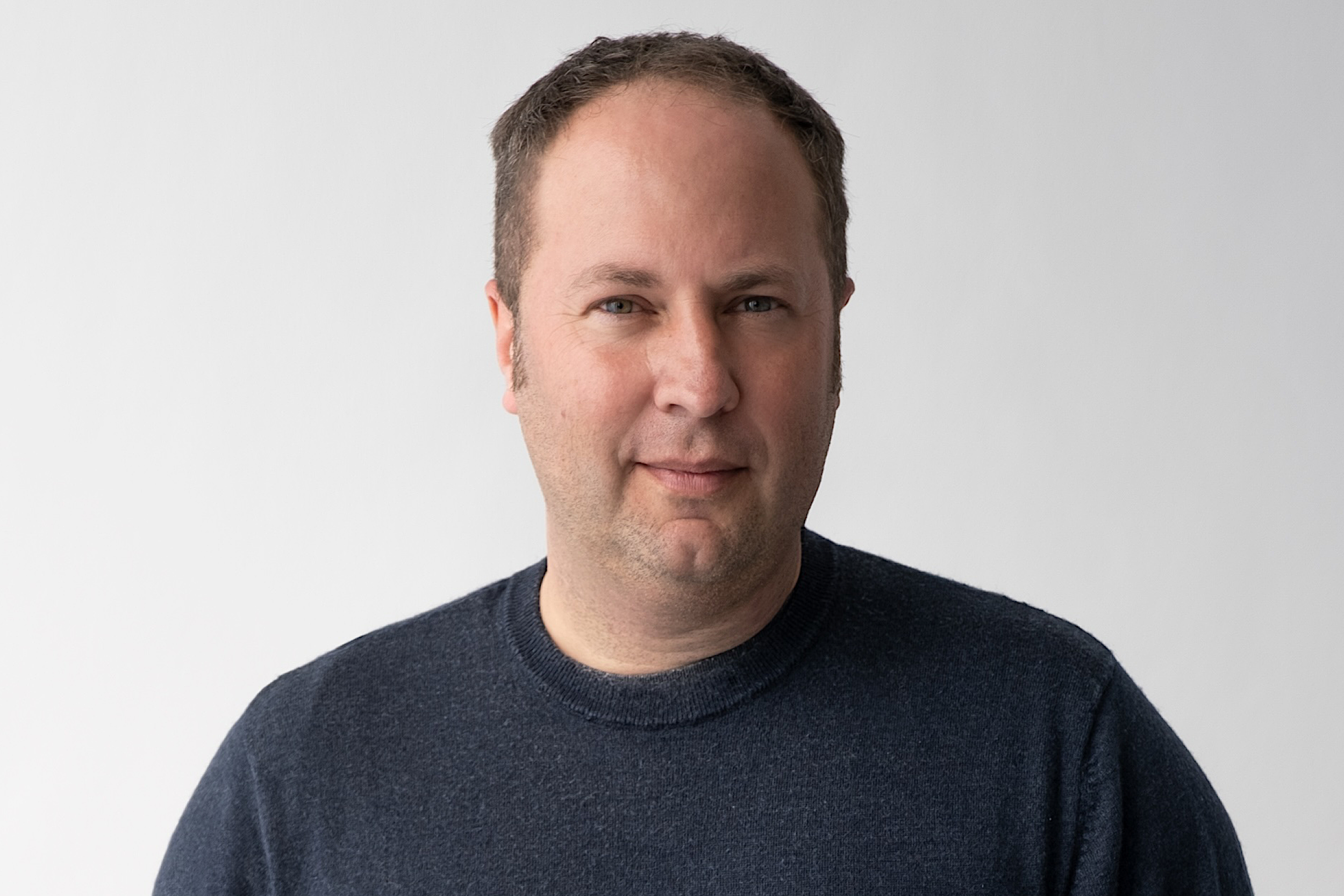 Porträt eines Mannes mit kurzem Haar und dunklem Pullover vor hellem Hintergrund.