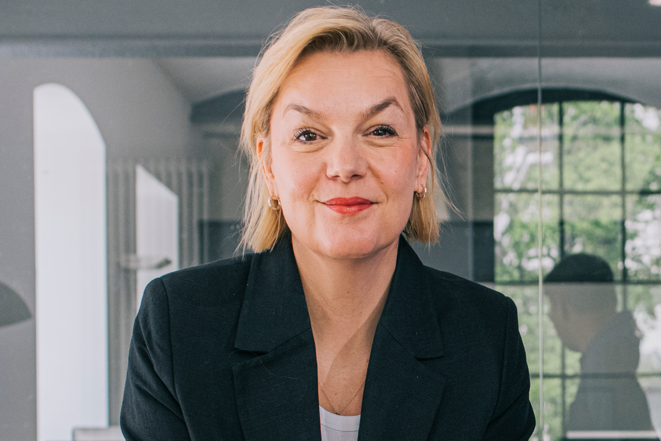 Porträt einer Frau mit blondem Haar und schwarzem Blazer, lächelnd vor hellem Innenraum mit Fenster.