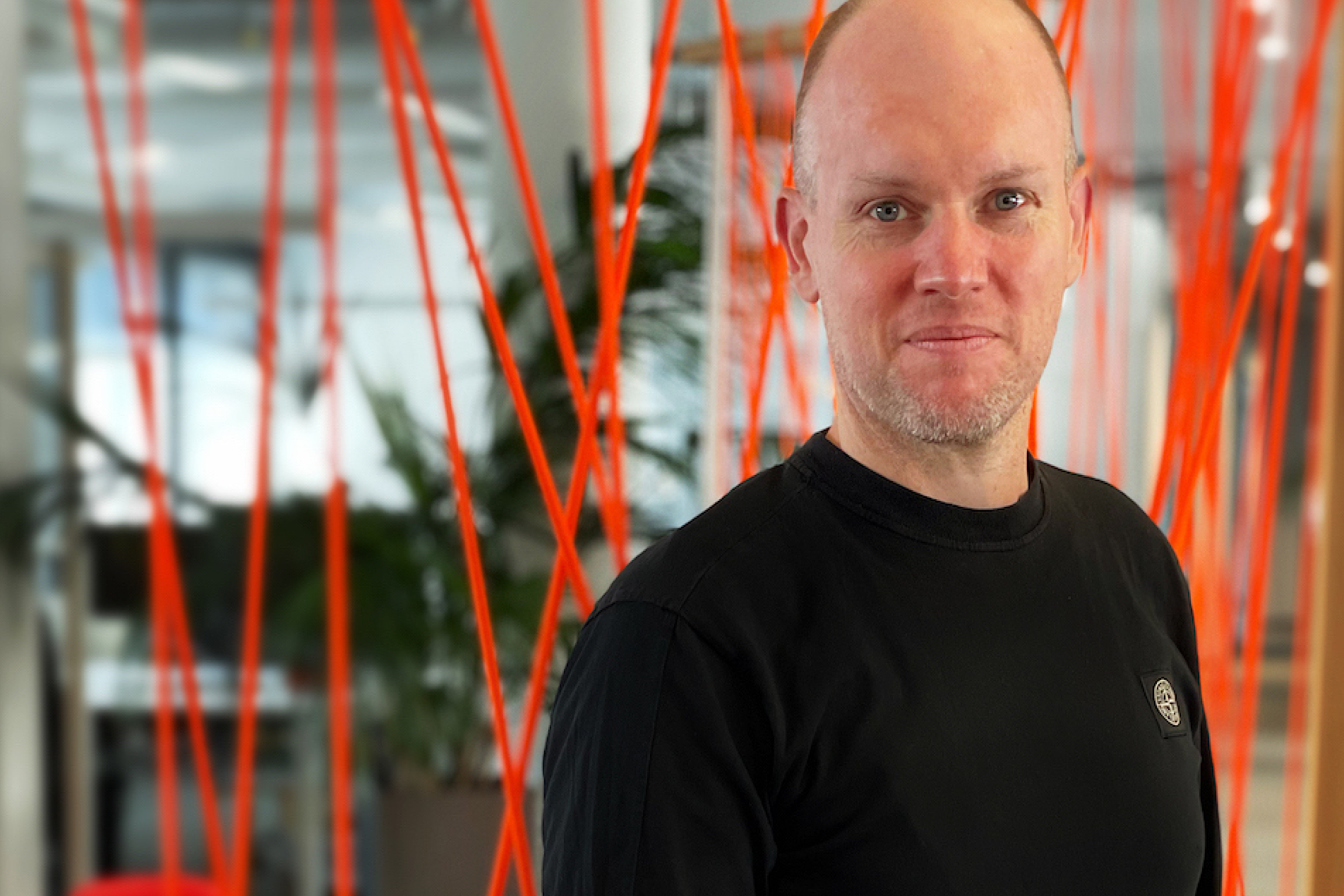 Porträt eines Mannes mit rasiertem Kopf und schwarzem Shirt vor Bürohintergrund mit roten Linien.