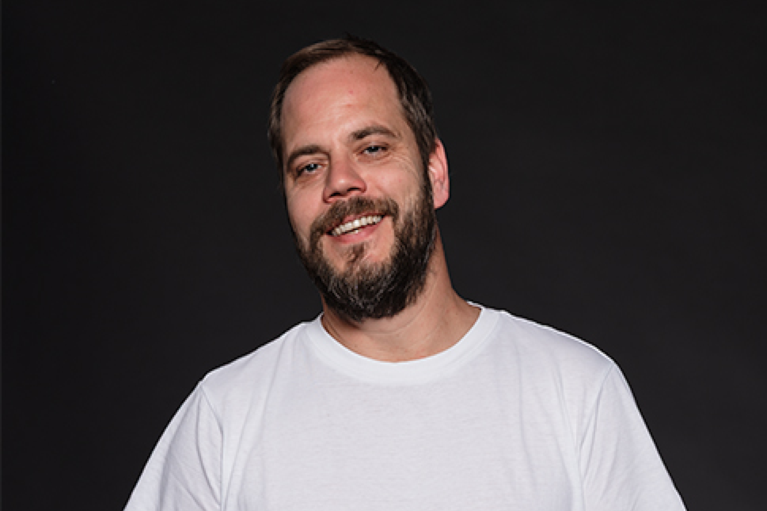 Porträt eines lächelnden Mannes mit Bart und weißem T-Shirt vor dunklem Hintergrund.